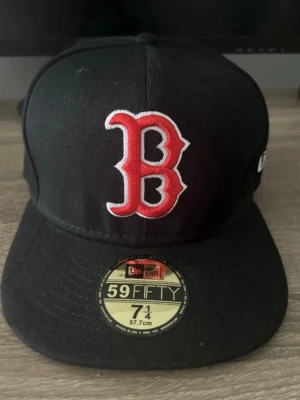 Svart Boston Red Sox keps 59FIFTY - Snygg svart keps från New Era 59FIFTY med Boston Red Sox logga i rött och vitt framtill. Klassisk flat brim och broderad detalj på sidan. Tillverkad i bomull och polyester, storlek 7 1/4 (57,7 cm). Perfekt för dig som gillar sportig streetstyle.