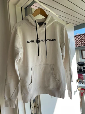 Vit hoodie från Sail Racing - Snygg vit hoodie från Sail Racing med svart tryck på bröstet och svarta dragsnören i huvan. Klassisk känguruficka framtill och mjukt material som känns skönt mot huden. Perfekt för dig som gillar stilren och sportig look. Bokstäverna är dock lite slitna!