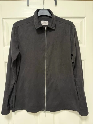 Svart overshirt med dragkedja ”zipjacka” - Stilren svart overshirt med hel dragkedja framtill och klassisk krage. Tillverkad i ett mjukt tyg som ger en avslappnad look. Perfekt att bära över en t-shirt eller skjorta för en enkel men snygg outfit. Använd men bra skick. 