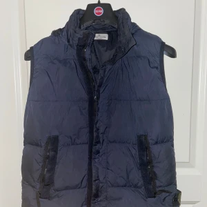 Stone Island Väst Marinblå - Säljer denna snygga Stone Island väst eftersom jag vuxit ur den. Modellen heter Stone Island Junior Nylon Down Vest och har utfällbar luva. Nypris ligger runt 4000kr. Skick 8/10. Jag hittar ingen lapp som säger storleken men jag uppskattar att den sitter som XS om inte lite större! västen är äkthetsverifierad genom certilogo.
