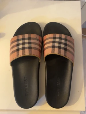 Rutiga slides från Burberry - Snygga slides från Burberry med klassiskt beige, svart och rött rutmönster på remmen. Sulan är svart och har en enkel, stilren design. Perfekta för sommaren och ger en lyxig touch till din outfit.