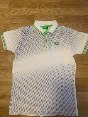 Vit prickig piké från Hugo Boss/ Som S storlek stretchiga  - Säljer en fräsch vit pikétröja från Hugo Boss med små svarta prickar och gröna detaljer. Kragen har gröna ränder och loggan är broderad i grönt på bröstet. Perfekt för dig som gillar sportig och stilren look.