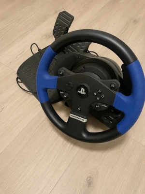 Thrustmaster ratt och pedaler PS4/PS5 - Säljer en Thrustmaster ratt och pedaler till PlayStation. Ratten har svart och blått grepp, flera knappar och PlayStation-logga i mitten. Perfekt för racing och bilspel, kopplas enkelt in med sladd. Tillverkad i plast och gummi för bra känsla.