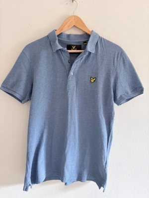 Blå pikétröja från Lyle & Scott - Klassisk blå pikétröja från Lyle & Scott med gul logga på bröstet. Tröjan har krage, korta ärmar och knäppning med två knappar framtill. Tillverkad i mjuk bomull som känns skön mot huden. Perfekt till en avslappnad och stilren look. Tröjan är nästan oanvänd och i mycket fint skick och färgen är perfekt.