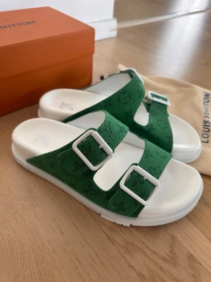 Gröna Louis Vuitton sandaler - Snygga gröna sandaler från Louis Vuitton med två breda remmar i tyg och vita spännen. Sulan är vit och har en chunky design. Remmarna har diskret LV-monogrammönster som ger en exklusiv känsla. Perfekta för dig som vill ha något trendigt och bekvämt.