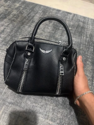Svart handväska Zadig & Voltaire - Snygg svart handväska i grained läder från Zadig & Voltaire. Väskan har silverfärgade detaljer, dubbla handtag och dekorativa dragkedjor framtill. Prickad med den ikoniska vingloggan i metall. Perfekt storlek för det viktigaste och ger en cool vibe till din outfit.