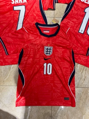 England Bellingham matchtröja röd - Säljer en röd England fotbollströja från Nike med nummer 10 och Bellingham på ryggen. Tröjan har svarta och vita detaljer, broderat landslagsmärke och stjärna på bröstet. Perfekt för dig som vill stötta England på match eller träning.