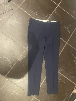 Mörkblå kostymbyxor H&M Slim Fit - Snygga mörkblå kostymbyxor från H&M i modellen Slim Fit. Byxorna har klassisk design med pressveck, bälteshällor och smal passform. Perfekta för en stilren och modern look. Storlek EUR 50 (US 34R).