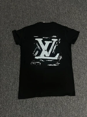 Svart Louis Vuitton t-shirt - Snygg svart t-shirt från Louis Vuitton med stort LV-tryck på ryggen och logga framtill i vitt. T-shirten har rund hals och korta ärmar. Perfekt för dig som vill ha en stilren men ändå statement look.