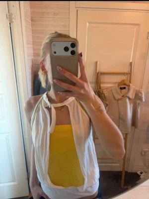 Vit och gul halterneck topp - Säljer en snygg halterneck topp med vitt ytterlager och gul insida. Toppen har knytning i nacken och ärmlös design, vilket ger en cool och somrig vibe. Materialet ser ut att vara mjukt och stretchigt, perfekt för varma dagar.