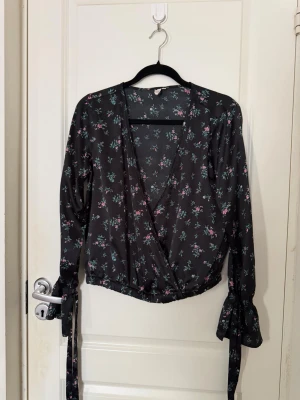 Blommig omlottblus från NLY Trend - Snygg svart omlottblus från NLY Trend med småblommigt mönster i rosa, grönt och vitt. Blusen har långa ärmar med knytband vid handlederna och resår i midjan för en feminin siluett. Perfekt till jeans eller kjol för en trendig look.