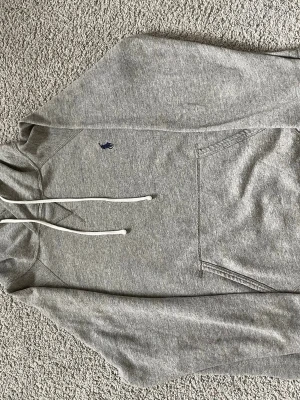 Grå Polo Ralph Lauren hoodie  - Snygg grå hoodie med klassisk huva och vita dragsnören. Har en magficka framtill och en liten mörkblå logga broderad på bröstet. Tillverkad i mjukt sweatshirtmaterial som är skönt att ha på sig. Perfekt för chill dagar eller när du vill ha en enkel och stilren look. Passar till allt! Endast en liten fläck (se bild). Storlek =medium men passar även Small. Priset kan diskuteras. Hör av er vid frågor!