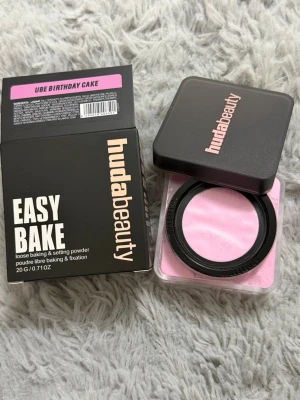 Huda Beauty Easy Bake Ube Birthday Cake - Lös baking- och settingpuder från Huda Beauty i färgen Ube Birthday Cake. Pudret har en ljusrosa ton och kommer i en fyrkantig plastburk med svart lock. Perfekt för att fixera makeup och ge en jämn finish.