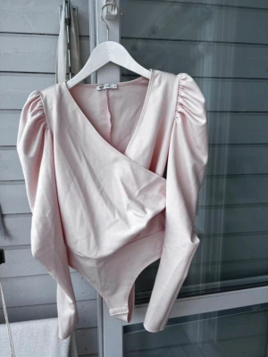 Ljusrosa body med puffärmar - Trendig ljusrosa body med långa puffärmar och omlott-ringning. Tillverkad i ett mjukt, glansigt material som ger en lyxig känsla. Perfekt för dig som vill ha en stilren och modern look med extra volym på axlarna.