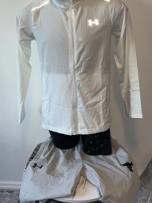 Vit vindjacka från Under Armour - Snygg vit vindjacka från Under Armour med hel dragkedja och huva. Jackan har två sidofickor och reflekterande logga på bröstet. Perfekt för blåsiga dagar och sportiga outfits. Lätt material som känns smidigt och bekvämt.