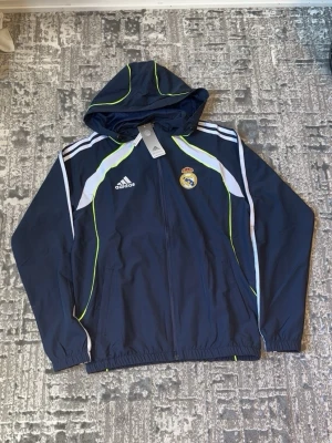 Mörkblå Real Madrid träningsset Adidas - Snyggt mörkblått träningsset från Adidas med Real Madrid-logga på både jacka och byxor. Setet har vita och neongula detaljer, huva på jackan och dragkedja framtill. Tillverkad i lätt och funktionellt material, perfekt för träning eller uppvärmning.