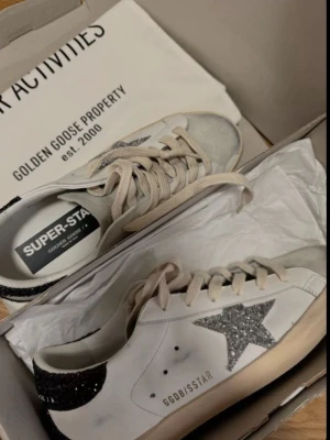 Golden Goose vita sneakers med glitter - Golden Goose sneakers i vitt läder med silverglittrig stjärna på sidan och svart glitterdetalj på hälen. Beige snören och slitna detaljer för en cool vintage-look. Modellen är låg och har GGDB/SSTAR-tryck på sidan.