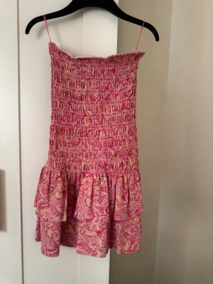 Rosa mönstrad kort klänning Gina Tricot - Supersöt kort klänning från Gina Tricot i rosa toner med smock upptill och volang nedtill. Klänningen har tunna axelband och ett färgglatt mönster i rosa och ljusa nyanser. Perfekt för varma dagar och sommarens alla tillfällen.
