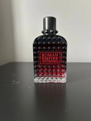 Roman Empire Intenso parfym valentino doup - Snygg parfymflaska med texten 'Roman Empire Intenso'. Flaskan är rektangulär med rundade kanter och har en cool design med nitar över hela ytan . Färgen går från svart upptill till röd och sedan klar nertill. Metallfärgad kork i toppen.