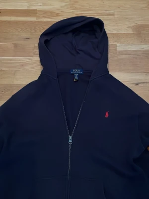 mörk Blå hoodie från Polo Ralph Lauren - Snygg mörkblå hoodie med dragkedja från Polo Ralph Lauren. Klassisk röd logga broderad på bröstet och stor huva. Tillverkad i mjukt bomullsmaterial och har fickor framtill. Perfekt för en avslappnad och stilren look.