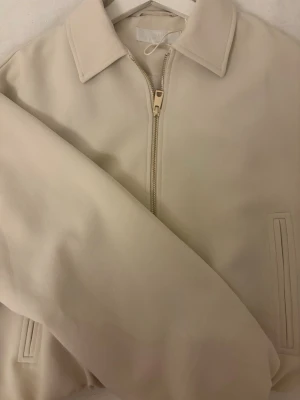 Ljusbeige bomberjacka med gulddragkedja(aldrig använd) - Stilren beige bomberjacka med klassisk krage och dragkedja framtill. Jackan har två diskreta fickor och ribbade muddar vid ärmslut och nederkant. Perfekt för en clean och modern look.