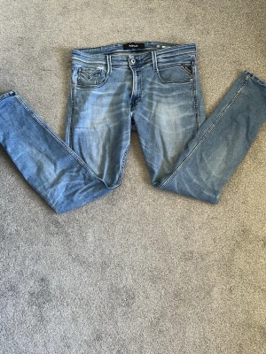 Blå slim fit jeans från Replay - Snygga blå slim fit jeans från Replay med klassisk femficksdesign och slitna detaljer. Jeansen har dragkedja och knapp i midjan samt dekorativa sömmar på bakfickorna. Perfekta för en avslappnad och trendig look. Det finns ett litet hål vid ena fickan men är inget större problem. Stl W 32 L 32