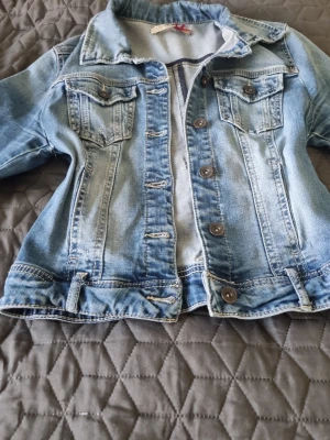 Klassisk blå jeansjacka - Snygg blå jeansjacka med slitna detaljer och bruna knappar. Jackan har två bröstfickor med lock och knapp samt markerad söm längs ryggen. passar till allt. I väldigt fin skick I storlek M kan passa s också