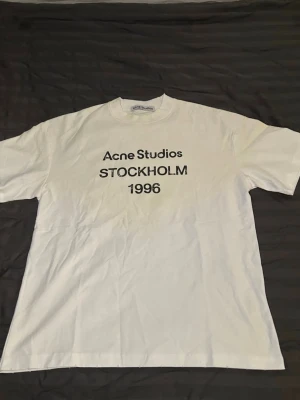 Acne Studios t-shirt - Säljer en vit t-shirt från Acne Studios med svart text 'STOCKHOLM 1996' på bröstet. Klassisk passform med rund hals och korta ärmar. Perfekt för dig som gillar stilrena och trendiga plagg.