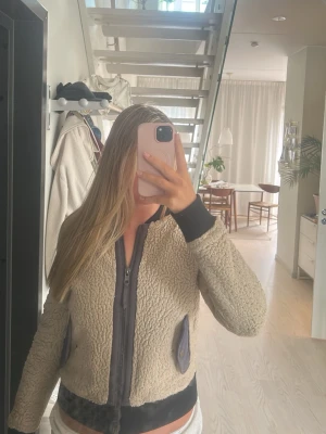Beige teddyjacka från Marrakech - Säljer en beige teddyjacka från Marrakech med svarta muddar och detaljer. Jackan har dragkedja framtill och två fickor med knappstängning. Perfekt för dig som vill ha en mysig och trendig jacka till hösten.