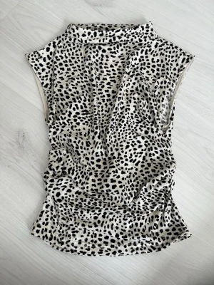 Leopardmönstrad ärmlös topp - Säljer en snygg ärmlös topp med leopardmönster i svart och beige. Toppen har en hög hals och är tillverkad i ett mjukt, stretchigt material som sitter bekvämt mot kroppen. Perfekt för dig som vill sticka ut lite extra.