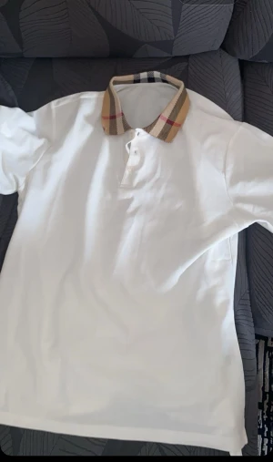Vit t-shirt med rutig krage - Snygg vit t-shirt med klassisk rutig krage i beige, svart och rött. T-shirten har korta ärmar och knäppning framtill vid halsen. Perfekt för dig som vill ha en stilren look med en twist.