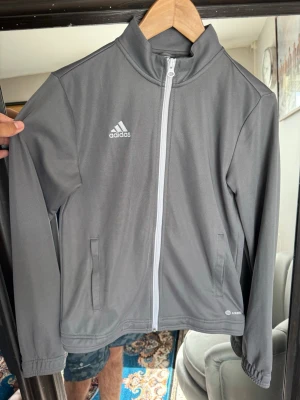 Grå Adidas träningsjacka AEROREADY - Snygg grå träningsjacka från Adidas med vit dragkedja och AEROREADY-teknologi. Jackan har hög krage, två sidofickor och Adidas-logga på bröstet. Perfekt för träning eller uppvärmning. Tillverkad i funktionsmaterial som andas.