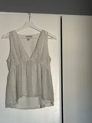 Vit prickig ärmlös blus - Söt vit blus med små svarta prickar och djup v-ringning. Modellen är ärmlös och har en lätt, luftig passform med något längre bakstycke. Perfekt för varma dagar och enkel att styla till olika looks.