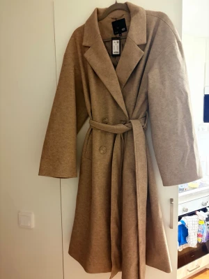 Beige kappa med knytskärp - Stilren beige kappa med bred krage och knytskärp i midjan. Kappan har dubbelknäppning framtill och raka ärmar. Perfekt för dig som gillar en klassisk och tidlös look. Passar till många olika outfits och ger en snygg siluett.