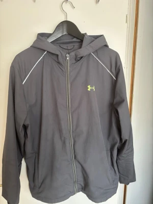 Grå vindjacka med huva från Under Armour - Snygg grå vindjacka från Under Armour med huva och hel dragkedja framtill. Jackan har vita detaljer vid axlarna och ett gult logotryck på bröstet samt stor logga på ryggen. Tillverkad i lätt och vindtätt material, perfekt för blåsiga dagar. Helt ny dragtjedja 