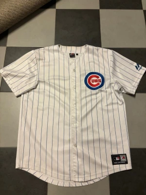 Chicago Cubs basebolltröja Majestic - Vit basebolltröja med blå ränder från Majestic, officiell Chicago Cubs med stort broderat Cubs-märke på bröstet. Knappar framtill och korta ärmar. MLB-logga nertill. Perfekt för sport och streetwear.