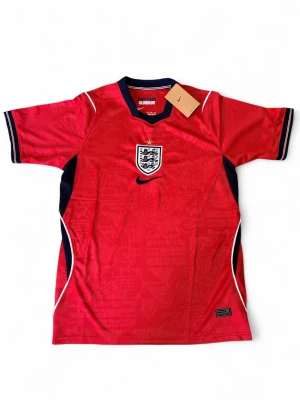 Röd England Nike fotbollströja - Röd England fotbollströja från Nike med broderat landslagsemblem och svart Nike-logga på bröstet. Tröjan har korta ärmar, marinblå och vita detaljer längs sidorna och halsen. Tillverkad i lätt och ventilerande material, perfekt för träning eller match.