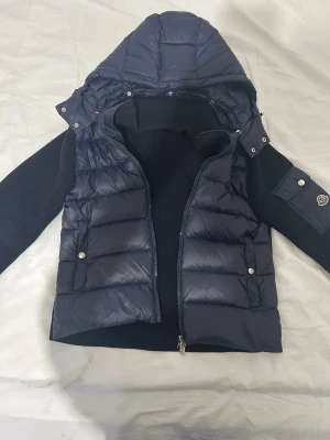 Mörkblå dunväst från Moncler - Snygg mörkblå dunjacka från Moncler med avtagbar huva och stickade ärmar. Jackan har dragkedja framtill, sidofickor med tryckknappar och en Moncler-logga på ärmen. Perfekt för lager-på-lager och streetstyle.
