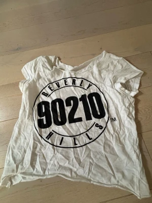Vit Beverly Hills 90210 t-shirt - Vit t-shirt med stort svart Beverly Hills 90210-tryck framtill. T-shirten har korta ärmar och coola slitsar i ryggen för en edgy look och den e lite  off shoulder har själv klippt i den!  Tröjan e använd mycket men fortfarande bra skick! Tröjan e xs men passar xxs 