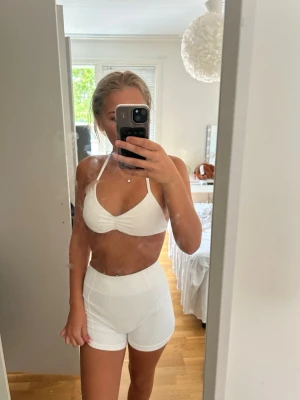 Vitt sportigt set med topp och shorts - Säljer ett vitt sportigt set med en halterneck topp och matchande höga shorts. Setet är sömlöst och har en tight passform, perfekt för träning eller chill. Materialet är stretchigt och mjukt för maximal komfort. Finns även scrunch i rumpan.