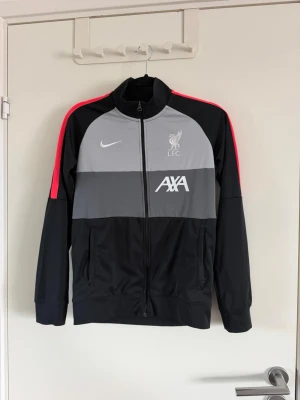 Liverpool Nike träningsjacka svart/grå - Snygg träningsjacka från Nike med Liverpool FC-logga och AXA-tryck. Jackan är svart med grå paneler och röda detaljer på axlarna. Hel dragkedja framtill, två fickor och hög krage. Perfekt för träning eller som sportig vardagsstil. Sparsamt använd.
