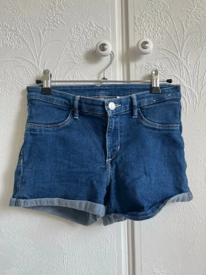 Blå denimshorts från H&M - Snygga blå denimshorts från &Denim med uppvikta benslut. Shortsen har normal passform, bälteshällor och stängs med knapp och dragkedja. Perfekta för sommardagar och enkla att matcha med olika toppar.