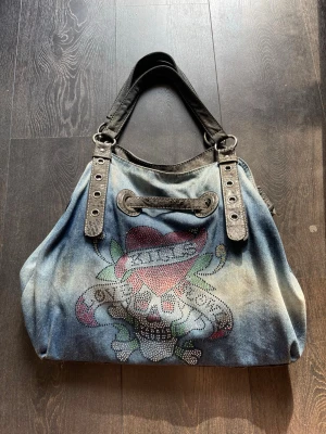 Ed hardy väska - Säljer en Ed Hardy väska då jag inte längre använder den. Enda egentliga slitningar är på handtagen. Supersnygg och har fått så mycket komplimanger för den här! Har 4 fack på insidan samt en där bak, väldigt rymlig!