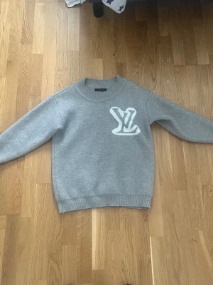Grå stickad fin tröja - Snygg grå stickad tröja med en stor vit -detalj både fram- och baksidan. Tröjan har rund halsringning, ribbade muddar och är gjord i ett mjukt stickat material. Perfekt för dig som gillar stilrena plagg med en twist.