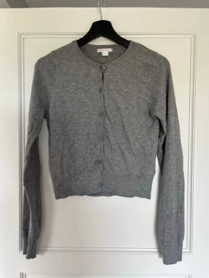 Grå kofta från Gina Tricot - Klassisk grå kofta från Gina Tricot med rund halsringning och knäppning framtill. Tillverkad i mjukt, tunt material som passar perfekt till lager på lager. Långa ärmar och enkel, stilren design som funkar till det mesta.
