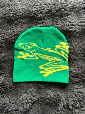Grön mössa med gul groda - Säljer en grön mössa med en stor gul groda som motiv på framsidan. Mössan är mjuk och stretchig, perfekt för att sticka ut i mängden. Passar dig som gillar färgglada accessoarer och vill ha något unikt till din stil.