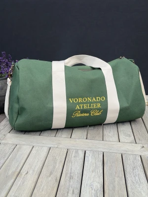 Voronado Riviera Duffelbag - Snygg grön duffelbag från Voronado Atelier med broderad gul text på sidan. Väskan har vita bärhandtag och axelrem i kraftig canvas, samt guldfärgade metalldetaljer. Perfekt för gymmet eller weekendresan, rymmer mycket och har en klassisk, sportig look.