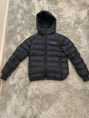  Peak Performance jacka  - Snygg marinblå pufferjacka från Peak Performance med huva och dragkedja framtill. Jackan har broderad logga på bröstet och två sidofickor med dragkedja. Perfekt för kalla vinterdagar och riktigt skön att bära.