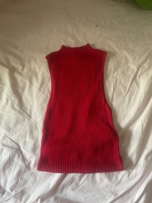 Ribbad röd ärmlös polotopp - Säljer en ribbad röd polotopp i stretchigt material. Toppen är ärmlös och har en hög hals, perfekt för att styla med jeans eller kjol. Snygg och enkel att matcha med olika accessoarer.
