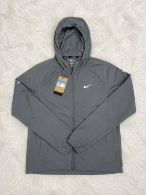 Nike miler grey - Snygg grå vindjacka från Nike med huva och dragkedja framtill. Jackan har en minimalistisk design med vit Nike-logga på bröstet och elastiska muddar vid ärmsluten. Perfekt för blåsiga dagar och enkel att matcha med olika outfits.
